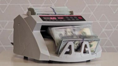 Bir para sayma makinesi hızla para yığınlarını işliyor, verimlilik ve hız gösteriyor. Görünür para düzgün düzenlenmiş dolar banknotlarını içeriyor..