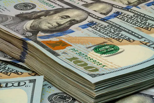 Düzenli bir şekilde yığılmış yüz dolarlık banknotların detaylı bir görüntüsü canlı renklerini ve çalışma alanındaki yumuşak ışıklandırma altında karmaşık tasarımlarını sergiliyor..