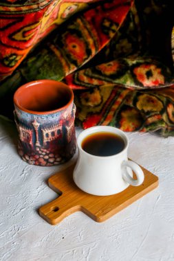 Küçük beyaz bir fincan renkli bir seramik kupanın yanında zengin kahve tutar. Dekor canlı, desenli bir masa örtüsüyle süslenmiş. Sahneye sıcaklık katıyor..