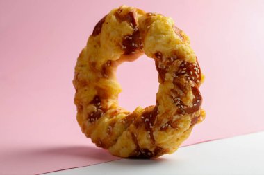 Yaratıcı şekilli bir donut yumuşak pembe bir arka plana karşı dik durur. Altın-kahverengi kabuğu susam tohumları ile tamamlanır. Çekici bir tasarım sergiler..