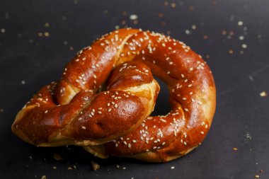 Sıcak, taze pişmiş susam tohumlarıyla kaplı kraker kırıntılarla dağılmış karanlık bir yüzeyde duruyor. Doku, altın-kahverengi kabuğunu ve yumuşak iç yüzünü sergiliyor..