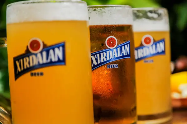 Farklı tipte biralarla dolu üç bardak yeşilliklerle çevrili ahşap bir masada otururken, insanlar açık havada güneşli bir kutlamanın keyfini çıkarıyorlar..