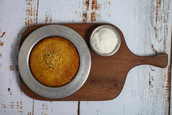Üzerine doğranmış fındık eklenmiş altın rengi bir tatlı, ahşap bir tahtanın üzerinde duruyor. Üstünde kremalı bir kase, davetkar bir hoşgörü..