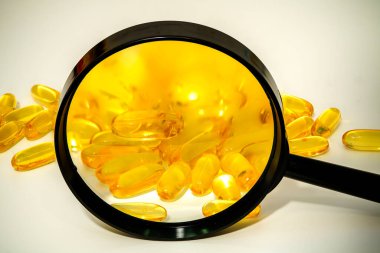 Close-up view features a magnifying glass highlighting yellow softgel capsules scattered on a white background, showcasing their smooth exterior and vibrant color. clipart