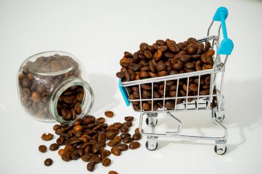 Bir alışveriş arabası kahve çekirdekleri ile doluyken, aynı koyu kavrulmuş fasulyelerle dolu açık bir kavanozun yanındaki beyaz bir yüzeye daha fazla fasulye dökülüyor..