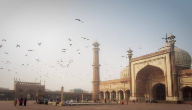 Yeni Delhi 'deki Jama Mescid Camii' nde gün batımında, güvercinler uçuşuyor ve bazı insanlar meydanda geziniyor..