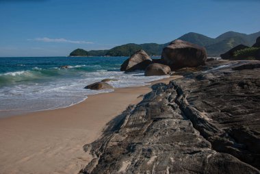 Kahverengi kumlu ve mavi suları olan bir sahil. Kayalarla dalgalı ve dağlarla ve yeşil ormanlarla dolu mavi bir arka plan.