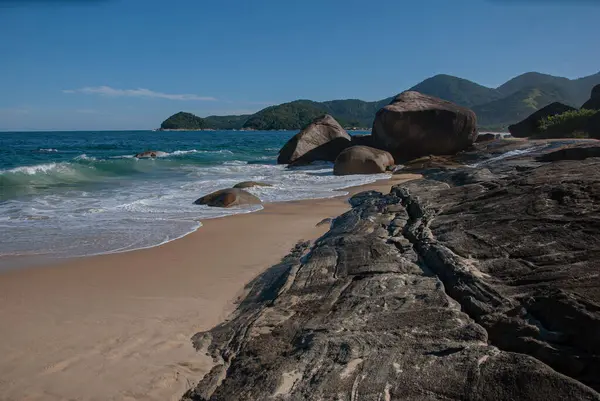 Kahverengi kumlu ve mavi suları olan bir sahil. Kayalarla dalgalı ve dağlarla ve yeşil ormanlarla dolu mavi bir arka plan.
