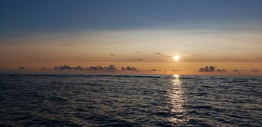 Gün doğumunda ya da günbatımında sakin bir deniz manzarası. Güneş ufukta batarken gökyüzünde ve denizde altın bir parıltı yayıyor. Sakin okyanus gökyüzü ile buluşmak için uzanır, alçak bulutların dağılımıyla