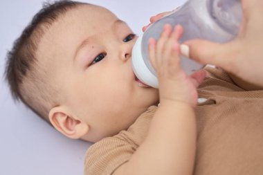 9 aylık çocuk yalan söylüyor ve bir şişe süt tutuyor, uykulu, beyaz arka planda ve doğal ışıkta yatıyor.