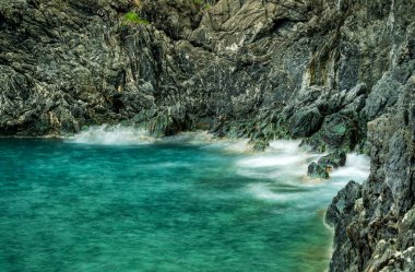 Net, turkuaz suların kayalara çarptığı sisli bir kıyı şeridi.