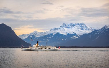 İsviçre Alplerinde gün batımında Cenevre Gölü 'nde bir tekne ve arka planda Les Dents du Midi, kar ve gökyüzünde bulutlar.