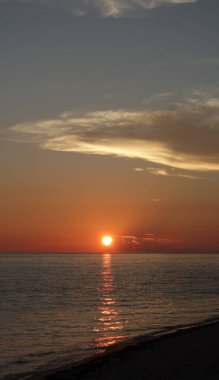 Suyun üzerinde parlak bir güneş yansıması olan gün batımı, yüksek, aydınlık bir bulutun yer aldığı dikey bir çerçeve içinde yakalanır.