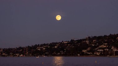 Mor gökyüzünde sarımsı bir dolunay karanın arkasından yükselir ve göllerin sularına ışıldar.