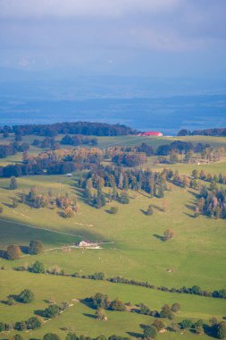 Uçsuz bucaksız yeşil bir manzara dalgalı tepeler, dağınık ağaçlar, uzak dağlar ve bulutlu bir gökyüzünün altında küçük evler. 