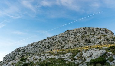 Berrak mavi gökyüzünün altında dağınık bitki örtüsüyle kayalık bir dağ. Arka planda uzak bir uçak izi var..