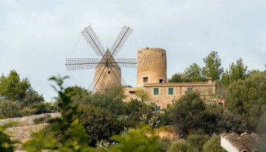 Yel değirmeni bir evin yanındaki tepede. Ev eski ve etrafında bir sürü ağaç var. Ön planda görünen tek şey yel değirmeni. Yatay fotoğraf