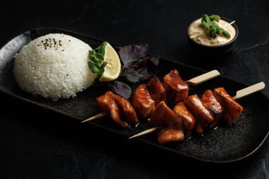 A plate of grilled salmon skewers glazed with teriyaki sauce, served alongside steamed white rice garnished with sesame seeds, a lemon wedge, fresh greens, and a side of creamy dipping sauce. clipart