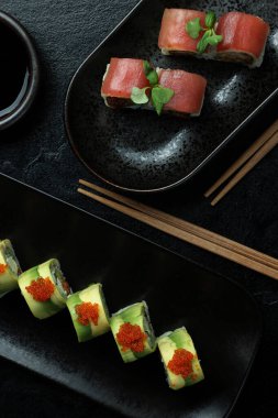 An elegant sushi platter featuring a variety of rolls on black plates with garnishes and chopsticks placed beside a soy sauce bowl. clipart