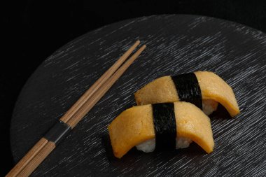 Tamago Nigiri featuring sweet Japanese omelet slices placed over sushi rice and tied with a strip of nori, presented elegantly on a dark plate with chopsticks. clipart