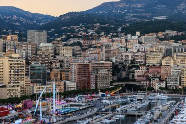 Monako, Monte Carlo - 01 Kasım 2024 - Parlak bir liman manzarası lüks yatlar, renkli eğlence parkı eğlenceleri, modern binalar ve parlayan günbatımı altında engebeli dağlar