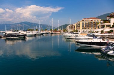 Karadağ 'ın Tivat kentinde zarif yatları ve sakin mavi sularıyla lüks bir marina. Modern binalar kıyı boyunca uzanır ve dağlar berrak bir gökyüzünün altında yükselir. Mükemmel bir Akdeniz manzarası.