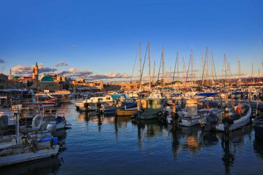 İsrail 'in Akko limanı altın gün batımında parlıyor. Gemiler ve yatlar sakin sularda dinleniyor, camiler ve açık mavi gökyüzü altındaki tahkimatlar ile tarihi şehri yansıtıyor..