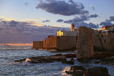 İsrail, Akko 'nun antik duvarları üzerinde çarpıcı bir günbatımı. Altın ışık deniz kenarındaki tarihi kaleyi aydınlatırken, dalgalar kayalık kıyılara çarpıyor. Barışçıl ama dramatik bir kıyı manzarası.