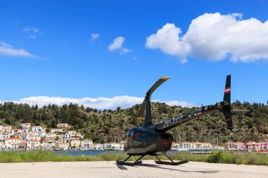 Yunanistan, Poros - Nisan 04, 2025 - Yunanistan 'ın Poros Adası' ndaki açık mavi gökyüzü altında, renkli evler ve yeşil tepeler manzaralı sahil kenarına park etmiş bir helikopter.