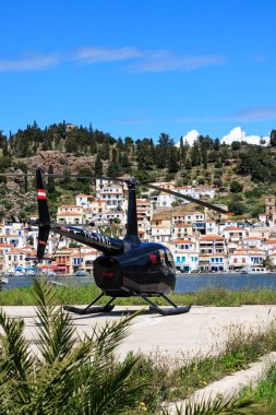 Yunanistan, Poros - Nisan 04, 2025 - Yunanistan 'ın Poros Adası' ndaki açık mavi gökyüzü altında, renkli evler ve yeşil tepeler manzaralı sahil kenarına park etmiş bir helikopter.