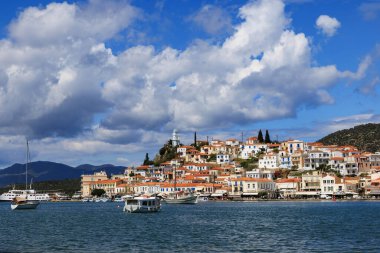 Poros Adası 'ndaki geleneksel Yunan şehirlerinde beyazlatılmış evler, tepe üstü bir saat kulesi ve dramatik bulutların altında sakin mavi bir körfeze demir atmış tekneler..