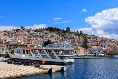 Yunanistan, Poros - Nisan 04, 2025 - Poros Adası limanında renkli yamaçlı evler ve arka planda açık mavi gökyüzü bulunan geleneksel Yunan feribotu.