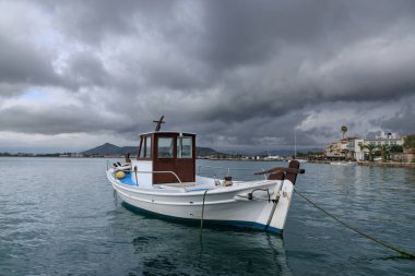 Yunanistan 'ın Nafplio kentindeki sakin denize demirlemiş beyaz ahşap balıkçı teknesi dramatik bulutlar ve uzak kıyı kasabasıyla.