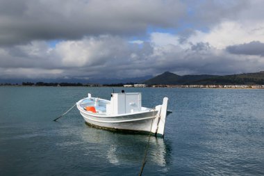 Yunanistan 'ın Nafplio kentindeki sakin denize demirlemiş beyaz ahşap balıkçı teknesi dramatik bulutlar ve uzak kıyı kasabasıyla.