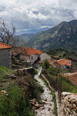 Arcadia, Moreloponnese 'deki Dimitsana' daki manzaralı dağ köyü yolu, taştan yolu, kırmızı kiremitli çatıları ve dramatik bulutlu gökyüzü altında yemyeşil tepeleri olan Yunanistan..