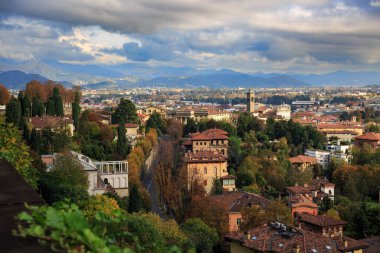 Bergamo, İtalya 'nın yüksek manzarası, terracotta çatıları, sonbahar yaprakları, bir kilise kulesi ve dramatik bulutların altındaki uzak dağlar..