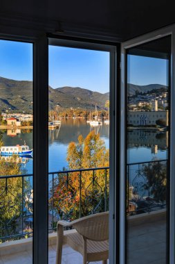 Yunanistan 'ın Poros kentindeki sakin deniz, tekne ve dağların manzarası, açık balkon kapılarından camlara yansıyan yansımalarla görülüyor.