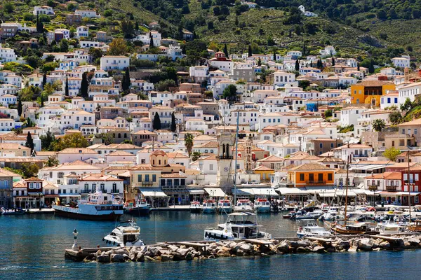 Yunanistan 'ın Hydra adasındaki büyüleyici liman kasabasının panoramik manzarası, beyazlatılmış evler, terasotta çatıları, tarihi bir saat kulesi ve güneşli bir günde limana yanaşan tekneler..