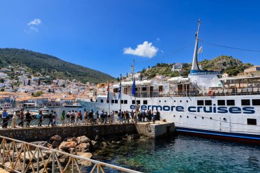 Yunanistan, Hydra - Nisan 06, 2025 - Yunanistan 'ın Hydra adasındaki bir Evermore Cruises gemisine binmek için liman boyunca yürüyen turistler. Kasabanın beyazlatılmış evleri ve terasotta çatıları arka planda canlı mavi gökyüzünün altındaki yamaca tırmanıyor.