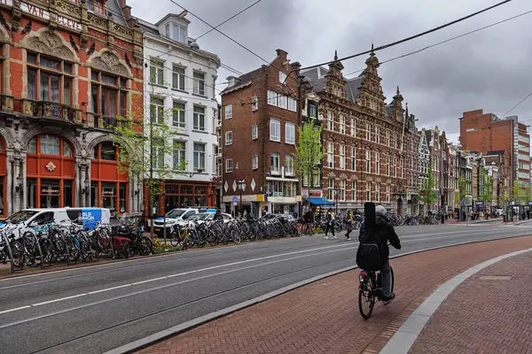 AMSTERDAM, NETHERLANDS - 11 Nisan 2024: Amsterdam 'da tarihi binalar, bisikletler ve bir bisikletçiyle dolu canlı bir cadde. Tramvay rayları yol boyunca kıvrılır ve insanlar bulutlu bir gökyüzünün altında kaldırımda yürür..