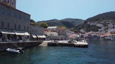 Yunanistan, Hydra - Nisan 06, 2025 - Yunanistan 'ın Hydra Adası limanının turkuaz suları, rıhtımlı tekneleri ve parlak Akdeniz gökyüzü altındaki geleneksel yamaç evleriyle canlı görüntüsü.