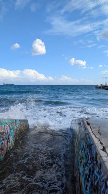 The vibrant blue water contrasts with the concrete walls adorned with colorful graffiti. In the distance, the horizon blends the sea and sky, with scattered white clouds adding depth to the blue sky clipart