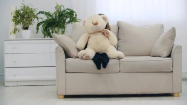 Kanepede oturan küçük gülümseyen kız oyuncak ayısıyla 4K yavaş çekim videosunda selam veriyor. Oyuncak ayılı kız kavramı.
