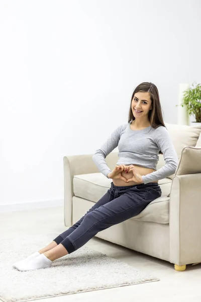 Photo of pregnant woman shows a heart shape on her stomach with her hands while sitting on the sofa in the living room and smiling at camera. Concept of pregnant woman