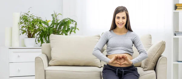 Photo of pregnant woman shows a heart shape on her stomach with her hands while sitting on the sofa in the living room and smiling at camera. Concept of pregnant woman