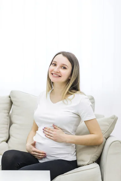 Photo of positive future mother sitting on white sofa. Concept of pregnant woman.
