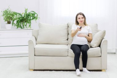 Pregnant woman sitting on sofa with remote control. Concept of pregnant woman with remote control.