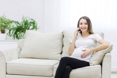 Smiling pregnant woman talking over the phone on white sofa. Concept of pregnant woman.