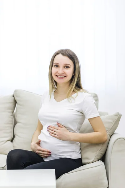 Photo of positive future mother sitting on white sofa. Concept of pregnant woman.