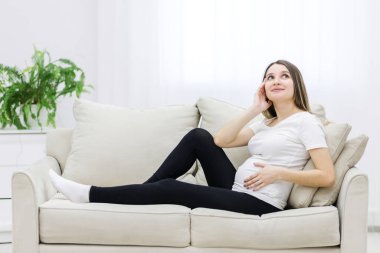 Young pregnant woman thinking about something on white sofa. Concept of pregnant woman.